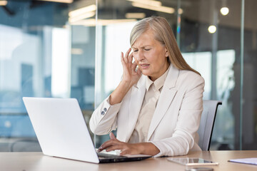 Confused senior business woman sitting in the office at the desk and holding her ear, feeling severe pain.