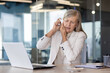 © Liubomir - Senior business woman sitting at the desk in the office and instilling medicine in her ears, grimacing in pain, suffering from ear inflammation.