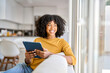 © insta_photos - Happy African American lady using tab device looking away on sofa at home. Smiling pretty young woman sitting on couch relaxing looking away at window holding digital table in kitchen.