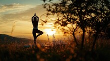  A Woman Doing Yoga In A Field With The Sun Setting Behind Her And A Tree In The Foreground, With The Sun Setting Behind Her, In The Distance.