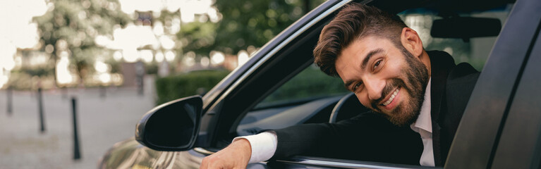 Smiling businessman in suit is riding behind steering wheel of car and looks camera