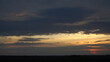 © Uldis - Panoramic view of a golden sunset sky with dramatic, dark grey clouds. The beautiful evening landscape showcases a visible horizon line on a plain at nightfall.