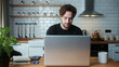 © mahirkart - Handsome curly hair man sitting in the kitchen using laptop enjoying surfing internet, using social media, chatting with friends, leaning back and smiling