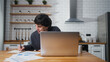 © mahirkart - Smiling young university student sitting in the kitchen at home, looking laptop computer, studying, taking notes. Male student researching, study at home.