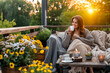 Young woman having a cup of tea on cozy wooden terrace with rustic wooden furniture, soft colorful pillows, light bulbs and flower pots. Charming sunny evening in summer garden.