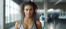 Tanned Young Student Girl In Sportswear Holds Bottle Looks At Camera With Wrapped Towel Around Neck At Fitness Club. Fit Hispanic Female At Break Of Exercise Of Fitness Training. Workout, Sport.