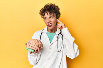 Wall Mural - Doctor holding a brain model on yellow studio covering ears with hands.