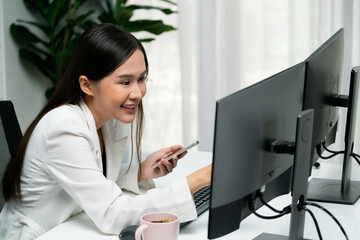 Smiling young Asian businesswoman searching marketing online strategy using smartphone to analyze digit report compare with pc in business analysis to make sales target in trader business. Stratagem.