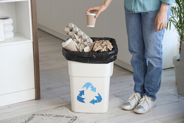 Poster - Woman throwing paper cup into full trash bin with recycle logo in kitchen