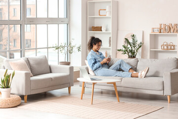 Wall Mural - Young African-American woman using mobile phone on sofa at home