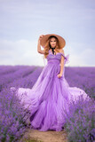 Fototapeta Kwiaty - beautiful woman in a lush lilac dress in a field of lavender. She's wearing a big braided hat