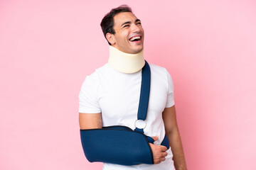 Wall Mural - Young caucasian man with broken arm and wearing a sling isolated on pink background laughing