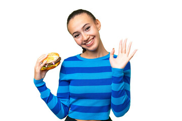 Canvas Print - Young Arab woman holding a burger over isolated background saluting with hand with happy expression