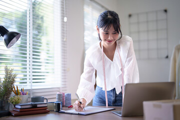 Young asian merchant women wearing measure tape in her neck and checking purchase online order on laptop to taking notes in clipboard while working about online shopping business in home office