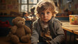 A curly-haired young child with a solemn gaze, seated next to a teddy bear in a classroom setting