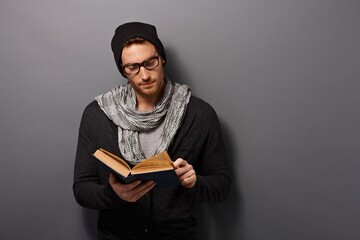 Poster - Happy urban style young man standing against grey wall, smiling, reading book, looking at camera.