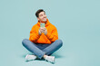 Full body young man he wears orange hoody casual clothes sitting hold in hand use mobile cell phone look aside isolated on plain pastel blue cyan color background studio portrait. Lifestyle concept.