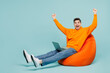 © ViDi Studio - Full body young IT man wears orange hoody casual clothes sit in bag chair hold use work on laptop pc computer do winner gesture isolated on plain pastel blue cyan color background. Lifestyle concept.