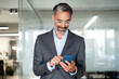 © Stock 4 You - Close-up of smiling mature Latin or Indian businessman holding smartphone in office. Middle aged manager using cell phone mobile app. Digital technology application and solutions for business concept.