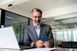 © Stock 4 You - Middle aged Hispanic business manager ceo using cell phone mobile app, laptop. Smiling Latin or Indian mature man businessman holding smartphone sit in office working online on gadget with copy space.