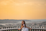 Fototapeta Krajobraz - Girl in a first communion dress celebrating her day on a viewpoint with the city and the bay in the background during sunset