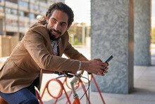Side view of mature Latin businessman with long hair using a smartphone while leaning on an orange bicycle, in a city environment