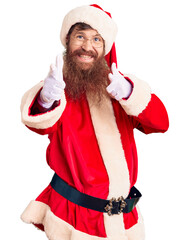 Sticker - Handsome young red head man with long beard wearing santa claus costume pointing fingers to camera with happy and funny face. good energy and vibes.