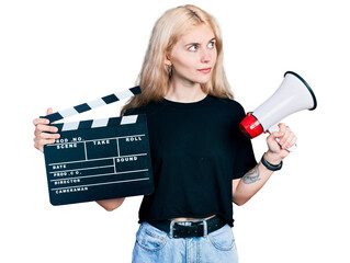 Canvas Print - Young caucasian woman holding video film clapboard and megaphone smiling looking to the side and staring away thinking.