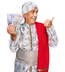 Canvas Print - Senior hispanic man wearing sherwani costume holding indian rupee banknotes pointing thumb up to the side smiling happy with open mouth