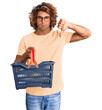 Young hispanic man holding supermarket shopping basket with angry face, negative sign showing dislike with thumbs down, rejection concept
