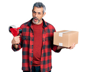 Wall Mural - Middle age hispanic with grey hair holding packing tape and cardboard box in shock face, looking skeptical and sarcastic, surprised with open mouth