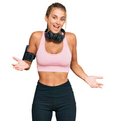 Wall Mural - Young blonde woman wearing gym clothes and using headphones clueless and confused expression with arms and hands raised. doubt concept.