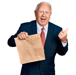 Wall Mural - Senior business man with grey hair holding take away paper bag pointing thumb up to the side smiling happy with open mouth