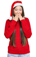 Wall Mural - Young chinese woman wearing christmas hat laughing and embarrassed giggle covering mouth with hands, gossip and scandal concept