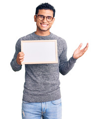 Wall Mural - Hispanic handsome young man holding blank empty banner celebrating victory with happy smile and winner expression with raised hands