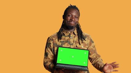 Wall Mural - Young guy showing pc with greenscreen layout on camera, pointing at isolated chromakey display. African american man presenting laptop with blank copyspace mockup screen in studio.