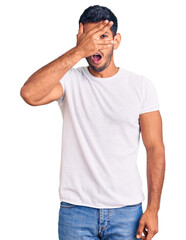 Sticker - Young latin man wearing casual clothes peeking in shock covering face and eyes with hand, looking through fingers with embarrassed expression.