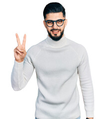 Wall Mural - Young arab man with beard wearing elegant turtleneck sweater and glasses showing and pointing up with fingers number two while smiling confident and happy.