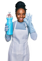 Poster - Young african american woman wearing apron holding detergent bottle doing ok sign with fingers, smiling friendly gesturing excellent symbol