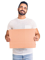 Wall Mural - Young hispanic man holding banner cardboard looking positive and happy standing and smiling with a confident smile showing teeth