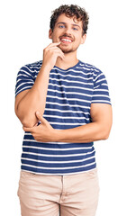 Wall Mural - Young handsome man with curly hair wearing casual clothes looking confident at the camera with smile with crossed arms and hand raised on chin. thinking positive.