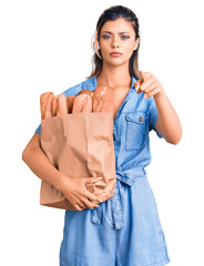 Sticker - Young beautiful woman holding paper bag with bread pointing with finger to the camera and to you, confident gesture looking serious