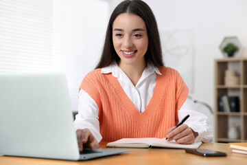 Canvas Print - Young woman writing in notebook while working on laptop at wooden table indoors