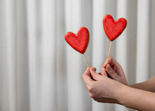 Hands Of Woman Holding Red Heart Shaped Cookies In Front Of White Curtain