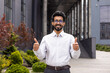 © Liubomir - Smiling young man in glasses and white shirt giving thumbs up outside modern office building, expressing approval and positivity.