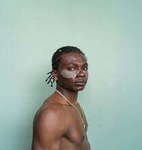 Side View Of Shirtless Black Man With Facial Cream On Wearing Earrings And A Necklace Stands Against A Light Green Backdrop