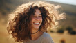 © red_orange_stock - Radiant young woman with voluminous curly hair smiling joyfully, bathed in the soft glow of sunlight outdoors.
