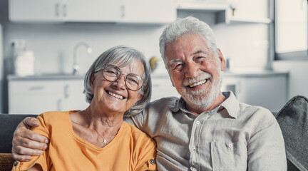 Wall Mural - Headshot portrait of smiling elderly 60s husband and wife sit relax on couch hugging cuddling, happy mature old couple rest on sofa in living room embrace look at camera show love and care.