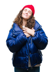 Sticker - Beautiful young brunette curly hair girl wearing winter coat, wool cap and sweater over isolated background smiling with hands on chest with closed eyes and grateful gesture on face. Health concept.