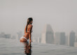 summer vacation travel photo - beautiful young perfect sexy body and skin wet girl in white bikini swimming and standing on the edge of pool on the top of hotel and looking in a city panorama